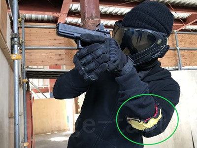 管理人の腕に巻いた黄色のマーカーを緑の丸で囲んだ写真(黒い服とゴーグルでハンドガンを構えたところ)