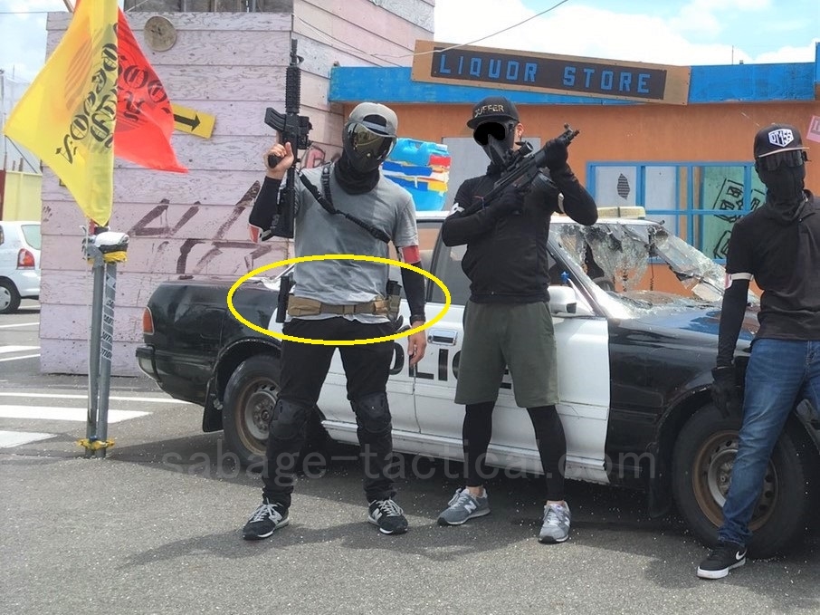 管理人のベルト装備を黄色の丸で囲んだ写真(パトカーの前に立つ管理人と友人たち)