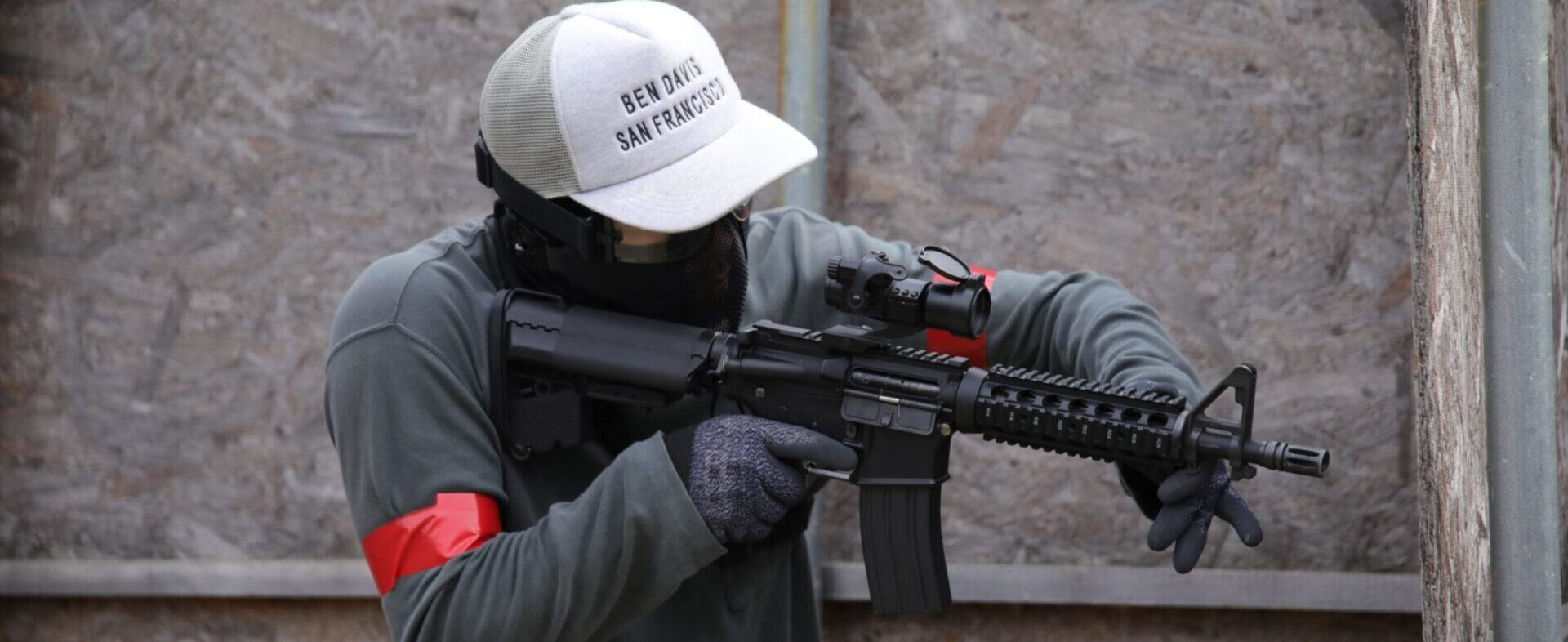 腕に赤いマーカーを巻き、キャップとマスクとゴーグルを着けて銃を構える管理人